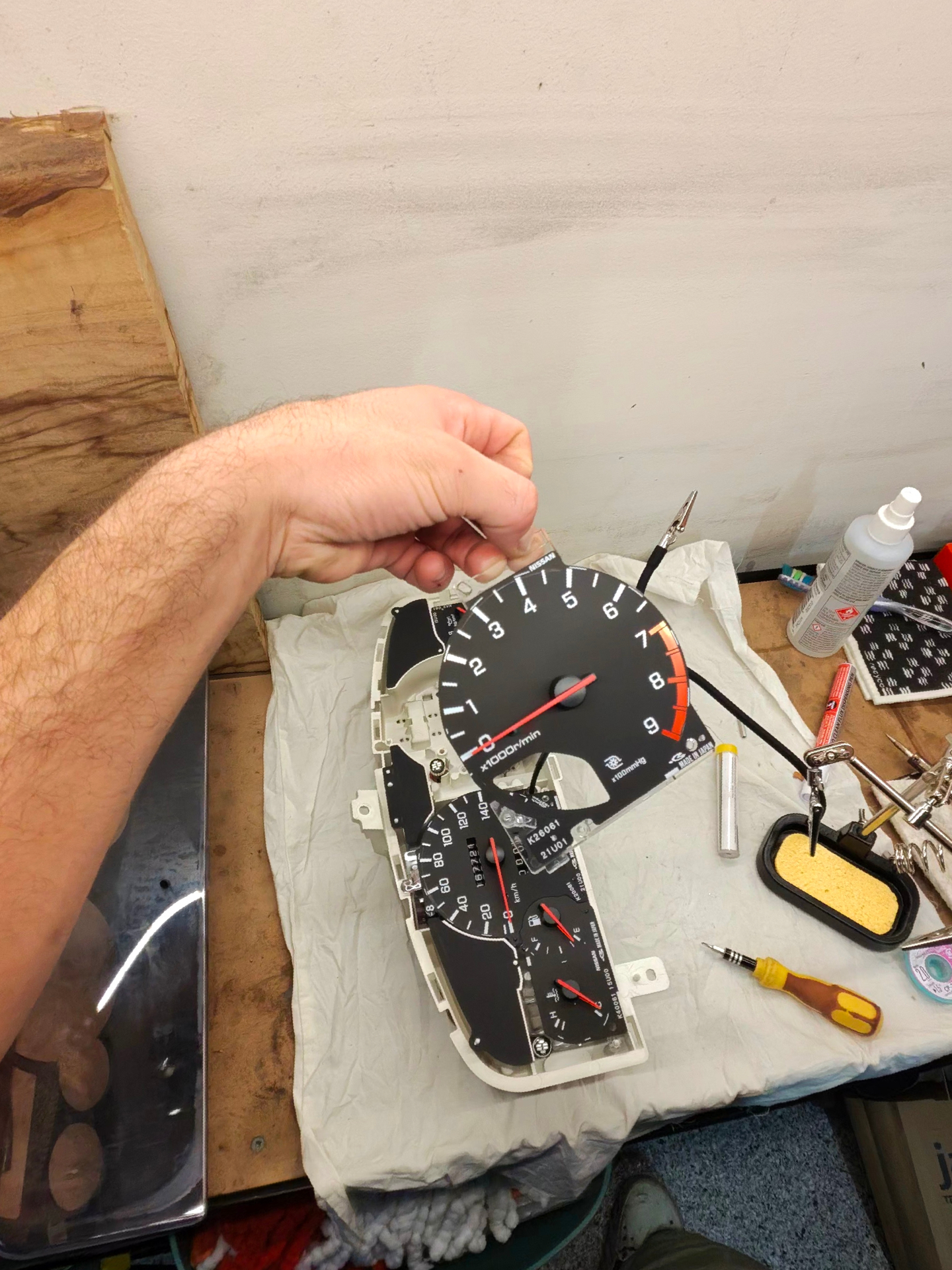 Hand lifting Nissan cluster dial face from housing on workbench with tools